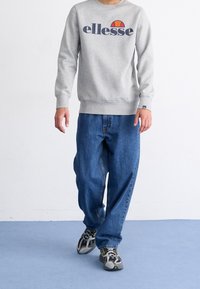 Grå sweatshirt med "ellesse"-logotyp, blå baggy jeans och svart/grå sneakers. Avslappnad outfit med räfflade manschetter och nederkant.