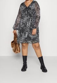 Vestido negro con estampado floral, mangas largas transparentes, escote en forma de V y un cinturón en la cintura. La modelo sostiene un bolso de piel sintética marrón, combinado con botas negras hasta la rodilla.