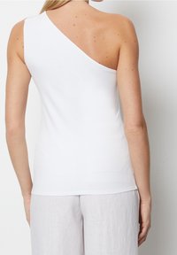 Haut blanc ajusté avec un décolleté asymétrique, en tissu lisse, dévoilant une épaule, associé à un pantalon de couleur claire, ample.