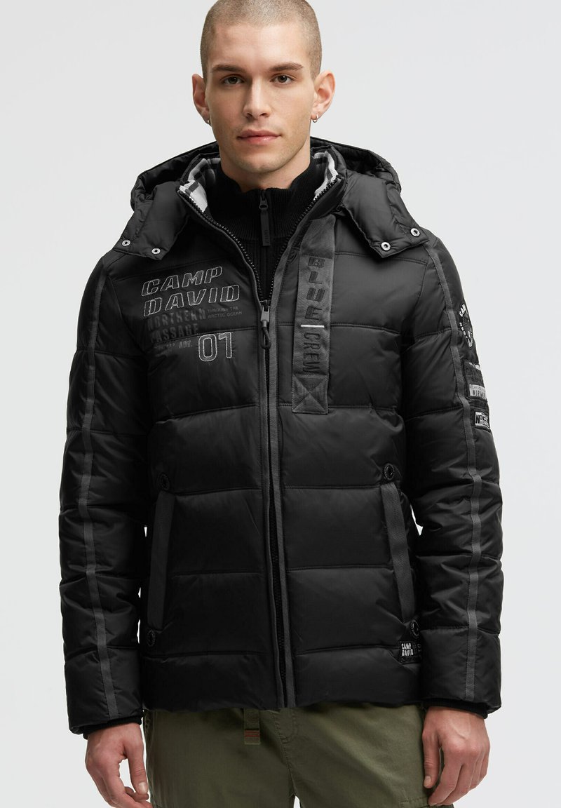 Schwarze gepolsterte Jacke mit Kapuze, ausgestattet mit gesticktem Text, Reißverschluss, Seitentaschen und grauen Akzenten an den Ärmeln. Strukturiertes und gestepptes Design.