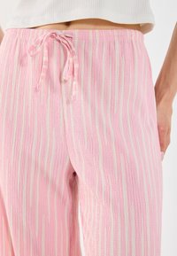 Różowe spodnie lounge z pionowymi białymi paskami, teksturowany materiał, elastyczny pas ze sznurkiem i luźny krój, podkreślające komfort i swobodny styl.