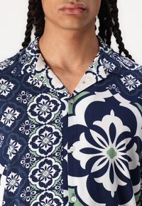 Camicia a maniche corte con un motivo floreale blu navy e dettagli bianchi e verdi, chiusura a bottoni e colletto a punta.