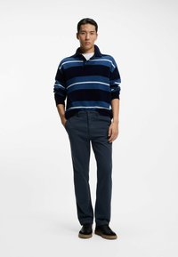 Marineblauer und blauer gestreifter Pullover mit Kragen, kombiniert mit dunkler Hose und schwarzen Schuhen. Der Pullover zeigt horizontale Streifen in unterschiedlichen Breiten.