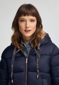 Granatowa kurtka puchowa z kapturem, z różowozłotymi zamkami błyskawicznymi i troczkami. Teksturowana powierzchnia, pikowany design oraz koszula dżinsowa pod spodem.