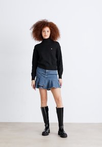 Melnā germanā turtlenecka džemperis, džinsu mini svārki ar kraukšķīgu malu un augstas līdz ceļiem spīdīgas melnas zābaki. Modelis stāv pret vienkāršu fona.