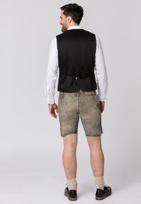 Schwarze Weste über einem weißen Hemd, kombiniert mit beigen Shorts. Die Weste hat ein Schnallendetail auf der Rückseite. Schwarze Schuhe und beige Socken runden den Look ab.