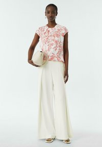 Ärmelloses Oberteil mit Blumenmuster in Weiß und Koralle, kombiniert mit weitem, cremefarbenem Hosen und einer beigen Handtasche. Riemchen-Heels runden den Look ab.