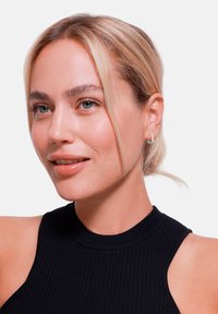 Modellen har på seg en svart ribbet topp uten ermer. Hun har blondt hår og er accessorert med små sølvøreringer, vist i profil.