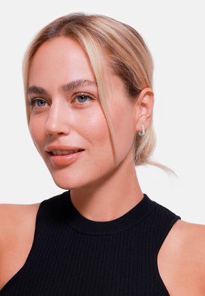 Modellen har på seg en svart ribbet topp uten ermer. Hun har blondt hår og er accessorert med små sølvøreringer, vist i profil.