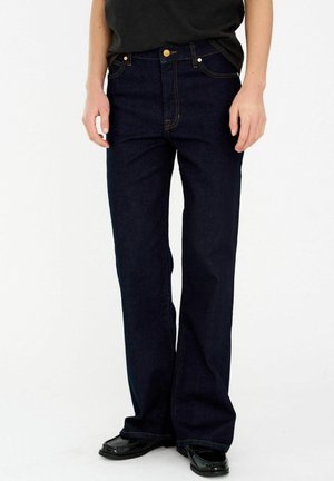 Persona que lleva jeans de pierna ancha azul oscuro con costuras a contraste, una camiseta negra y mocasines negros, parada contra un fondo blanco.