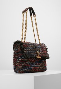 Tweedová kabelka s multikolorovým červeným, modrým a černým materiálem, zlatým řetízkovým ramenním popruhem a dekorativním zlatým uzávěrem na bílém podstavci.