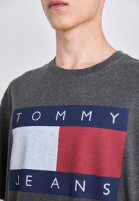 Camiseta de algodón gris con un logo central en bloques rectangulares azul, blanco y rojo, con el texto "TOMMY JEANS" en letras blancas en negrita.