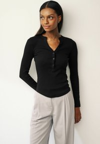 Black long-sleeve, ribbed henley top with a button placket and a rounded hem, paired with light gray pleated trousers.