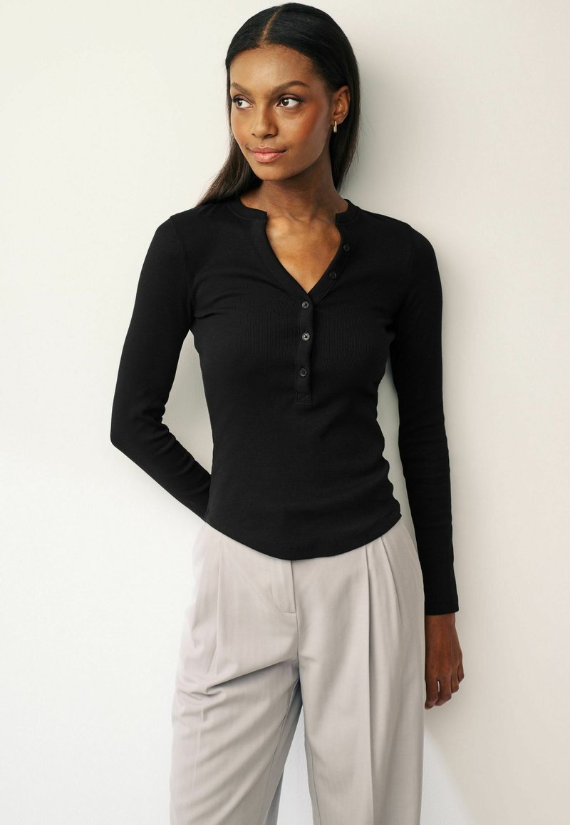 Black long-sleeve, ribbed henley top with a button placket and a rounded hem, paired with light gray pleated trousers.
