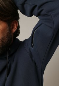 Navyblauwe sweatshirt met hoge kraag, ritszak op de linkermouw en koord bij de hals. Zachte, gestructureerde stof zichtbaar.