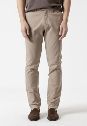 Hombre vistiendo pantalones chinos beige y mocasines de ante marrón, de pie con las manos relajadas al costado contra un fondo liso.