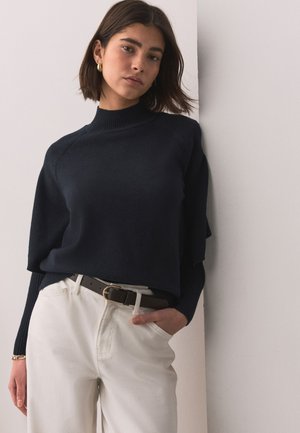 Frau mit kurzen braunen Haaren, die goldene Ohrringe trägt, schwarzen Rollkragenpullover und weiße Hose mit schwarzem Gürtel, steht vor einer weißen Wand.