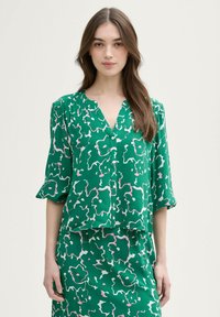 Grüne Bluse mit abstrakten weißen und pinken Blumenmustern, V-Ausschnitt, kurzen Ärmeln und weicher Textur. Kombiniert mit einem passenden Rock.