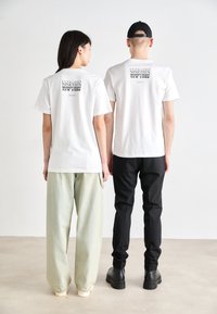 White cotton t-shirts with black print on the back, worn with loose light green and fitted black pants, demonstrating a casual look.