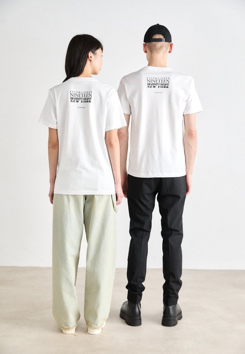 White cotton t-shirts with black print on the back, worn with loose light green and fitted black pants, demonstrating a casual look.