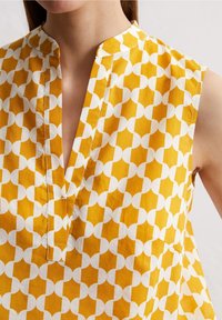 Blusa sin mangas amarilla y blanca con un patrón, cuello mandarín y escote en V, llevada por una persona, mostrando la parte superior del torso y el cuello.
