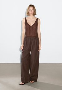 Hnědý lněný top bez rukávů s výstřihem do V a detaily na knoflíky, v kombinaci s hnědými lněnými kalhotami širokého střihu, které mají elastický pas a volný střih.