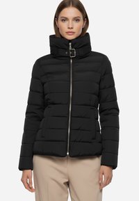 Giacca a vento nera con collo alto, cuciture orizzontali, zip frontale e tasche laterali. Texture liscia, design leggero e forma aderente.