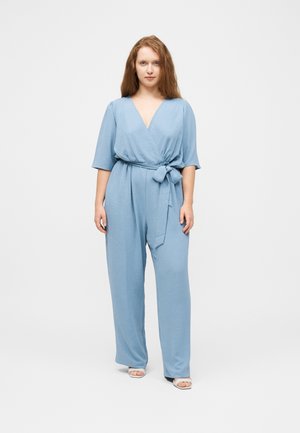 Nő áll, világoskék, átlapolt stílusú overált visel, derékban megkötve, és fehér, nyitott orrú magassarkú cipőt, egyszínű világos háttér előtt.