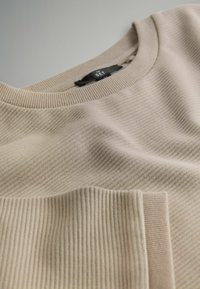 Beżowy sweter z prążkowanego dzianiny z okrągłym dekoltem i podwiniętymi rękawami, prezentujący teksturę i szczegóły tkaniny.