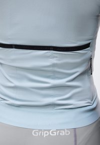 Κοντινό πλάνο της πίσω πλευράς μιας ανοιχτό μπλε μπλούζας ποδηλασίας με μια μαύρη οριζόντια τσέπη με φερμουάρ και το λογότυπο GripGrab πάνω από γκρι σορτς.