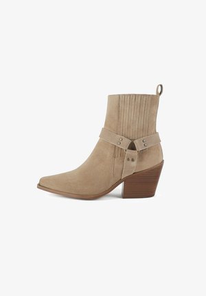 Beige suede ankle boot with a pointed toe, decorative stitching, and a leather strap with metal accents. Slightly raised wooden heel.