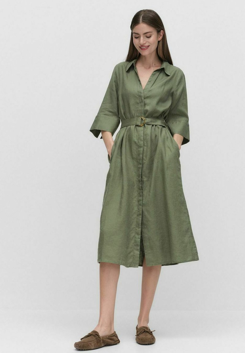 Zielona sukienka do kolan z kołnierzykiem, zapinana na guziki, z paskiem w talii i bocznymi kieszeniami; wykonana z lekkiego materiału o gładkiej fakturze.