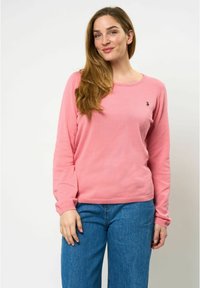 Roze langemouwen trui van zacht materiaal, met een ronde halslijn en een klein geborduurd logo op de borst. Gecombineerd met blauwe jeans.