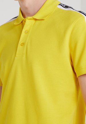 Polo jaune à manches courtes avec col et deux boutons, présentant des rayures à motifs blanc et noir sur les épaules.