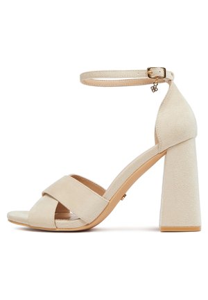 Beżowy zamszowy sandał na wysokim obcasie typu blok, z krzyżującymi się przednimi paskami oraz regulowanym paskiem wokół kostki z złotą klamrą i zawieszką.