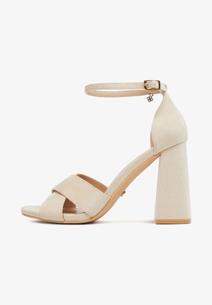 Sandalo beige in suede con tacco alto a blocco spesso, cinturini anteriori incrociati e cinturino alla caviglia regolabile con fibbia dorata e ciondolo.