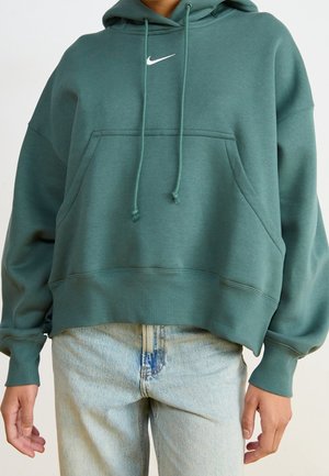 Person iført en blågrøn Nike-hættetrøje med snore og lyseblå jeans, stående med hænderne afslappet ned langs siderne mod en ensfarvet baggrund.