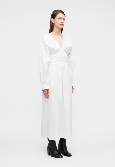 Γυναίκα που στέκεται, φορώντας ένα μακρύ λευκό φόρεμα με ραμμένη μέση και μανίκια, συνδυασμένο με μαύρα μποτάκια με τακούνι, σε λευκό φόντο.