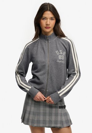 Kobieta nosząca szarą kurtkę na zamek z białymi paskami na rękawach i napisem "U.S.A 83 Superdry", w połączeniu z szarą plisowaną spódnicą w kratę z klamrą.