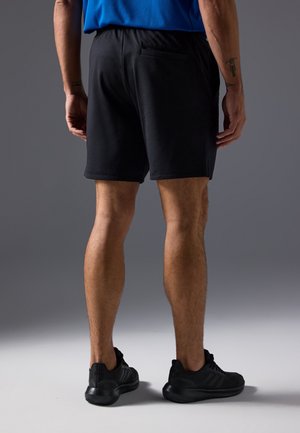 Mann trägt schwarze Sportshorts, schwarze Turnschuhe und ein blaues Shirt, steht mit dem Rücken zur Kamera vor grauem Hintergrund.