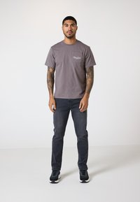 Grå bomull T-skjorte med rund hals, kortermede ermer og brystlomme med hvit tekst. Brukt med mørke jeans og svarte sneakers.