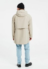 Giacca impermeabile beige con cappuccio, caratterizzata da un retro più lungo, fessure laterali e una vestibilità comoda. Abbinata a jeans azzurri chiari e stivali neri.
