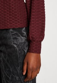 Burgunderfarbendes, strukturiertes Langarmoberteil mit einem gemusterten Design und gerafften Bündchen, kombiniert mit schwarzen Jeans. Fokus auf Details der Ärmel und des Stoffes.