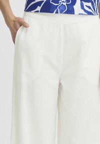 Pantalons larges blancs en tissu texturé, avec une taille élastique et des poches latérales. Le matériau semble léger et décontracté.