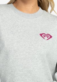 Sudadera gris con una textura suave, que presenta un pequeño diseño de corazón rosa en la parte izquierda del pecho. Cuello redondo y ajuste casual.