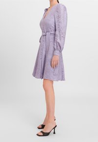 Lilac dress with a textured wave pattern, V-neckline, and puffed sleeves. Designed with a belt and a flared skirt. Black heeled sandals.