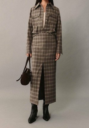 Personne portant une chemise à carreaux marron boutonnée, rentrée dans une jupe taille haute assortie avec une fente à l'avant, bottes noires et tenant un sac à main foncé.