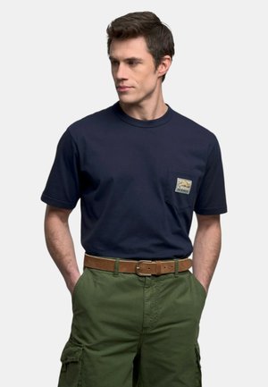 Uomo che indossa una t-shirt blu navy con logo sulla tasca sul petto e pantaloni cargo verde oliva con cintura marrone, in piedi con le mani in tasca.