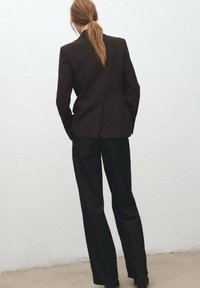 Czarna, dopasowana marynarka z pojedynczym rozcięciem z tyłu, zestawiona z czarnymi, prostymi spodniami. Gładka faktura, minimalistyczny design, bez widocznych elementów metalowych.
