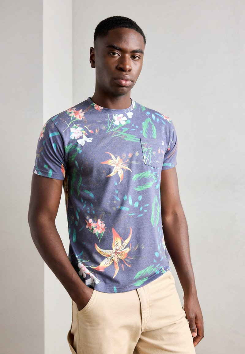 Camiseta de manga corta, ajustada, en tela azul oscura con un patrón floral en colores brillantes, que presenta un pequeño bolsillo en el pecho. Textura suave.